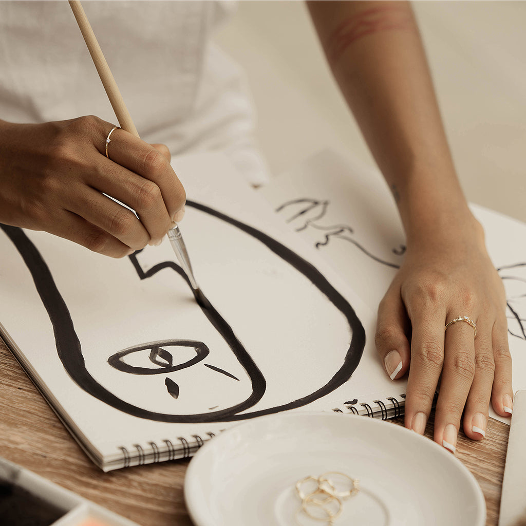A woman painting a face and wearing Ili ring and Kasai ring from Sceona fine jewellery